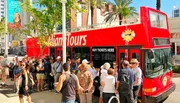 A group of people are gathered around a red double-decker tour bus, possibly waiting to buy tickets or board, on a sunny day in an urban area.