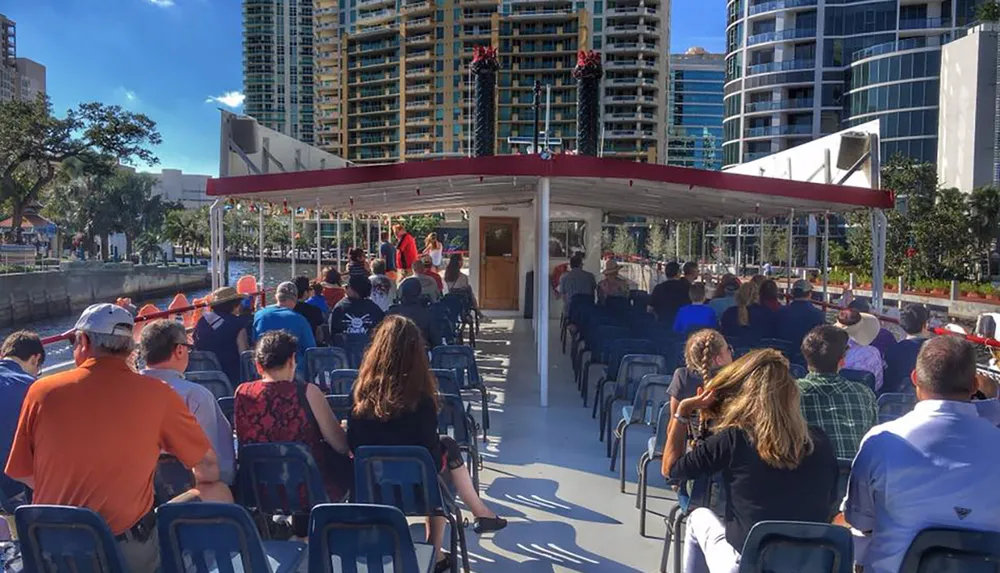 Passengers are seated on a tour boat with red awnings preparing to embark on a sightseeing tour amidst a backdrop of urban high-rise buildings on a sunny day