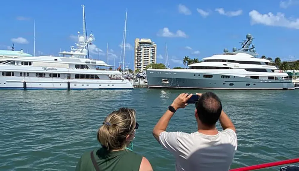 Two people are observing and photographing luxury yachts docked in a marina on a sunny day