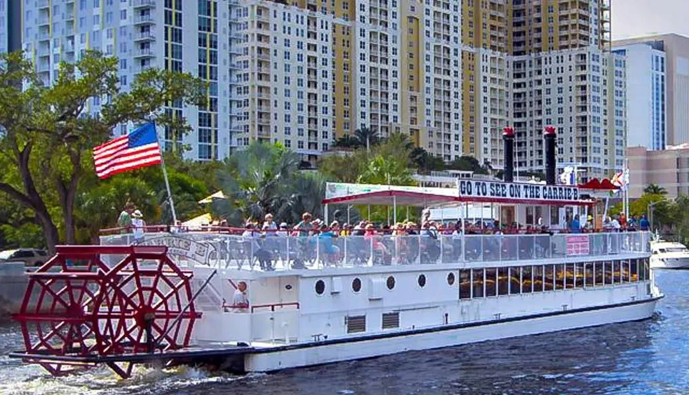 A paddlewheel riverboat filled with passengers is cruising on a river with an American flag flying in the air and a cityscape in the background