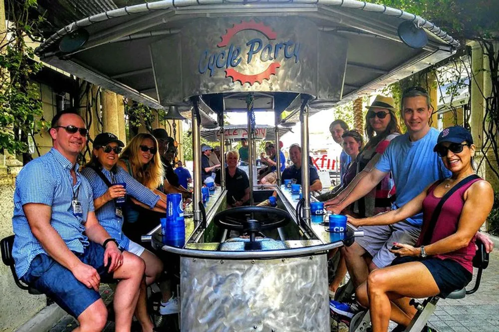 A group of smiling people are enjoying a pedal-powered bar on wheels known as a party bike while holding drinks