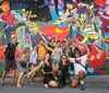 A group of cheerful people is posing with various playful gestures in front of a colorful and artistic mural with animated characters and floral designs