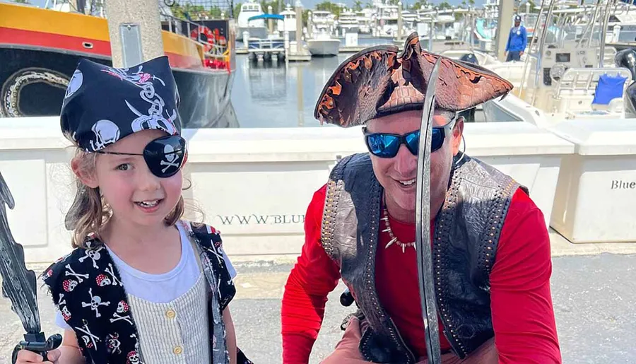A child and an adult dressed in pirate costumes are smiling with a marina in the background.