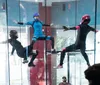 Three individuals in full-body suits and helmets are indoors skydiving in a vertical wind tunnel with an onlooker visible in the background