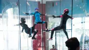 Three individuals in full-body suits and helmets are indoors skydiving in a vertical wind tunnel, with an onlooker visible in the background.