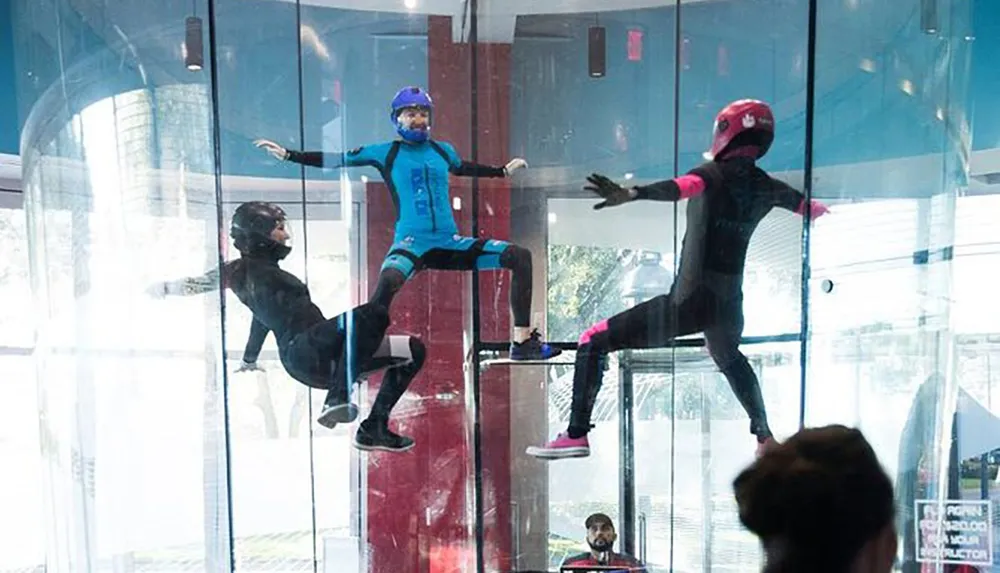 Three individuals in full-body suits and helmets are indoors skydiving in a vertical wind tunnel with an onlooker visible in the background