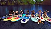 A group of cheerful people are posing on paddleboards on a calm water surface, surrounded by natural scenery.