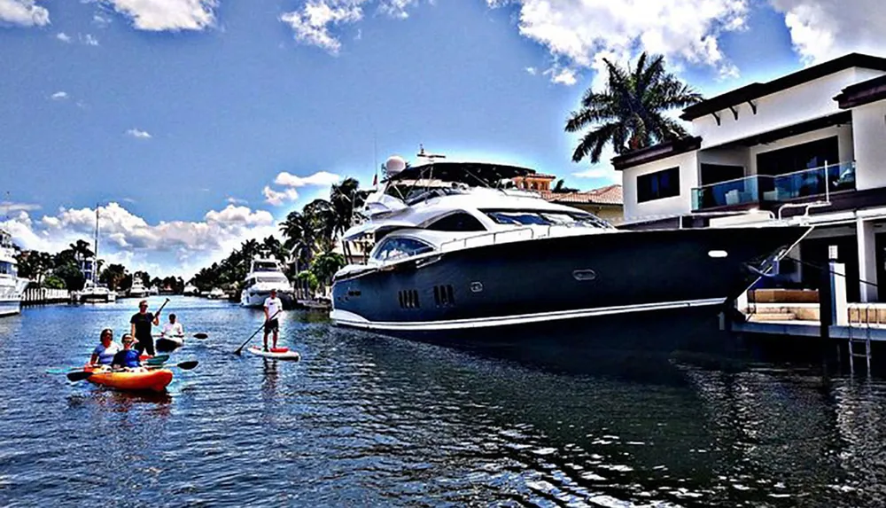 People enjoy water activities in a canal with a luxury yacht moored outside a waterfront home on a sunny day