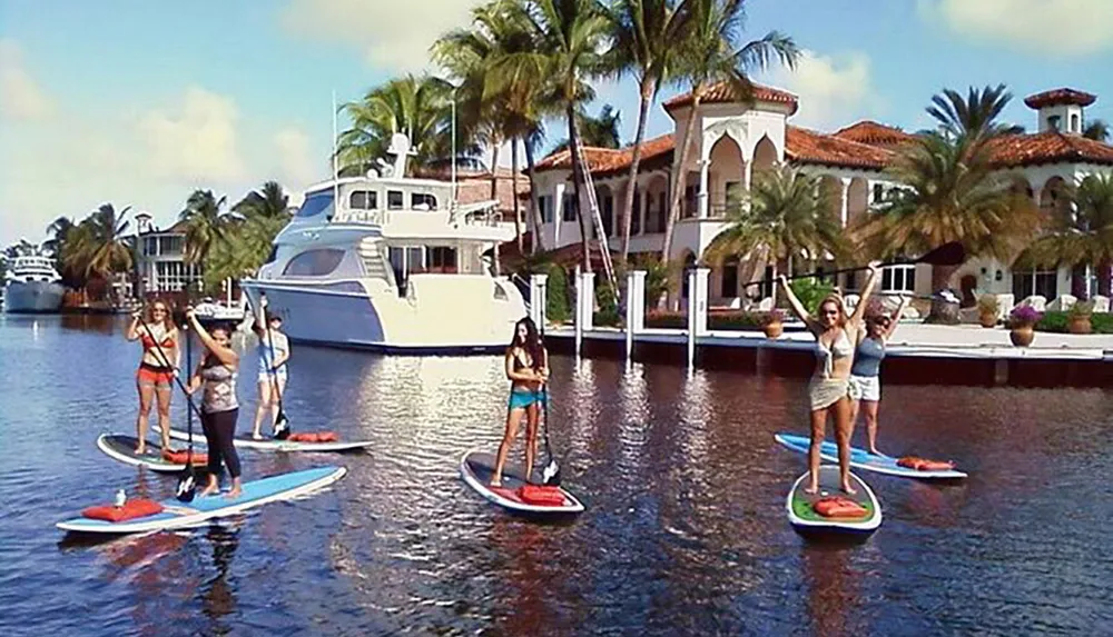 A group of individuals are paddleboarding in a calm waterway with a luxury yacht and waterfront homes in the background