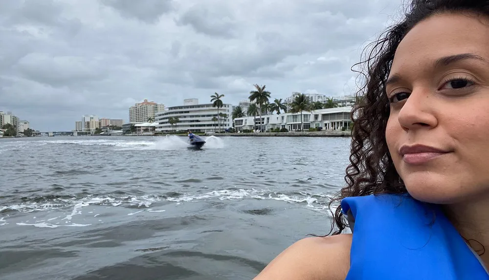 A person with curly hair wearing a blue life jacket is taking a selfie with a background of a waterway where another individual is riding a personal watercraft with waterfront buildings in the distance under a cloudy sky