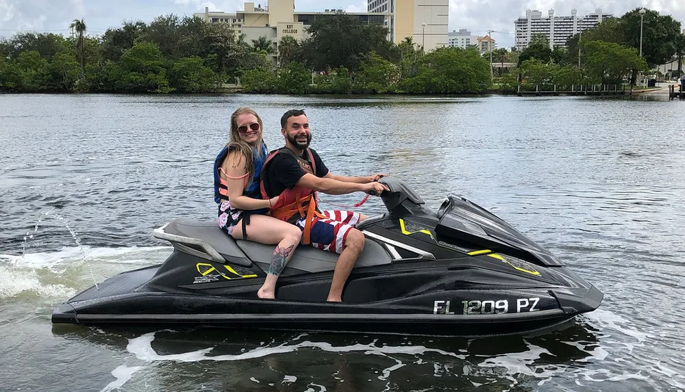 A man and a woman are smiling while sitting on a jet ski on a calm river with greenery and buildings in the background