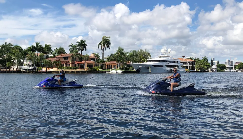 Two people are riding jet skis on a calm waterway with luxury homes and a yacht in the background