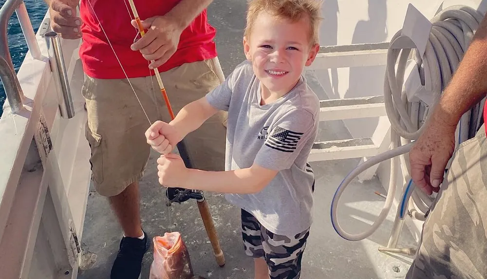 A smiling young child is proudly holding a fishing rod on a boat with two adults partially visible suggesting a family fishing trip