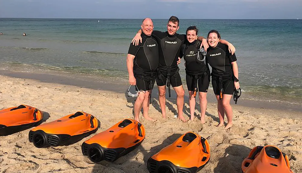 Four people dressed in black wetsuits with the brand HEAD visible are standing close together smiling on a sandy beach with four orange underwater scooters lying in the foreground and a clear blue sea in the background