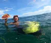 A person with diving goggles is smiling in the ocean while holding up a large shell and floating with the assistance of a bright lime-green swim board and a red buoy nearby