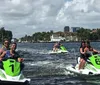 Three pairs of people are riding jet skis on a waterway with waterfront houses and buildings in the background