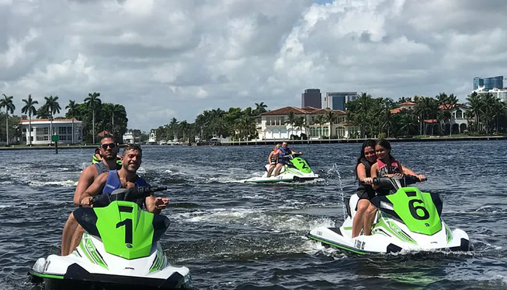 Three pairs of people are riding jet skis on a waterway with waterfront houses and buildings in the background