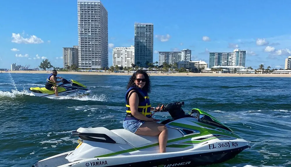 Two people are enjoying a sunny day riding personal watercraft near a coastline lined with buildings