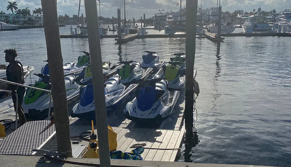 The image shows a person standing by a dock with multiple jet skis overlooking a marina under a clear sky