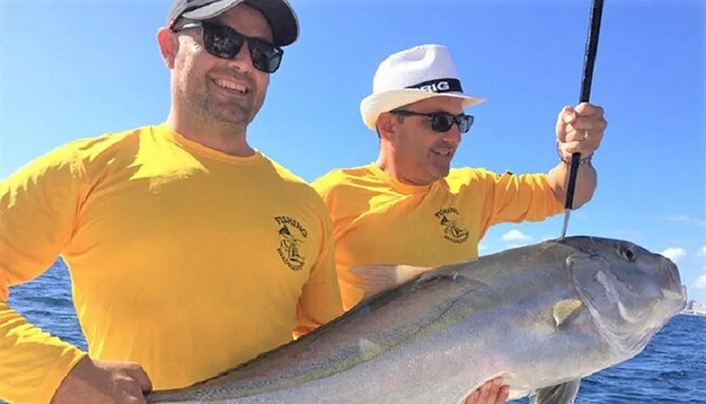 Two individuals in matching yellow shirts and sunglasses are smiling and posing with a large fish they caught aboard a boat on a sunny day