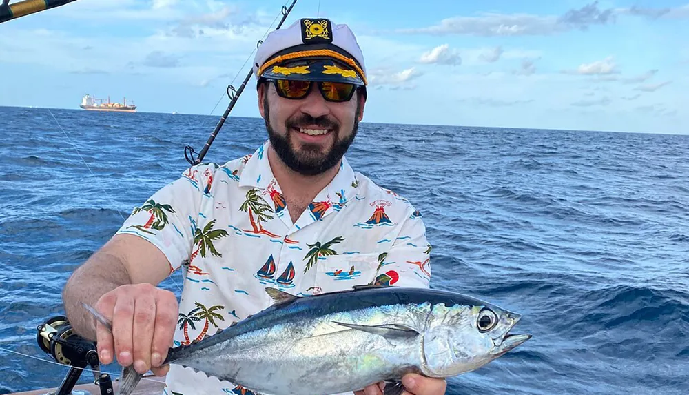 A smiling person in a captains hat and tropical shirt is proudly holding a freshly caught fish on a boat with the ocean and a ship visible in the background