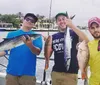 Three men are standing on a boat each proudly displaying their catch of fish with the water and some waterfront properties in the background