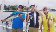 Three men are standing on a boat, each proudly displaying their catch of fish, with the water and some waterfront properties in the background.