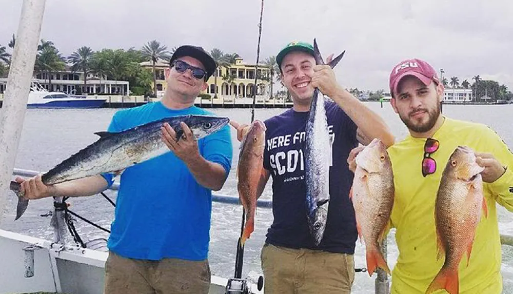 Three men are standing on a boat each proudly displaying their catch of fish with the water and some waterfront properties in the background