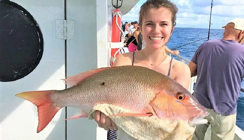 A smiling person is holding a large pinkish fish on a boat with other individuals in the background