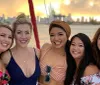 Five smiling women pose closely together for a photograph with a blurry sunset and city skyline in the background suggesting they are enjoying a social outing