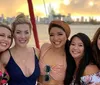 Five smiling women pose closely together for a photograph with a blurry sunset and city skyline in the background suggesting they are enjoying a social outing