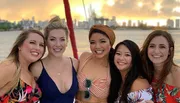 Five smiling women pose closely together for a photograph, with a blurry sunset and city skyline in the background, suggesting they are enjoying a social outing.