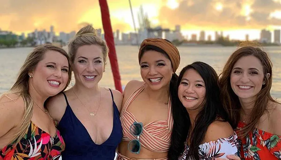 Five smiling women pose closely together for a photograph, with a blurry sunset and city skyline in the background, suggesting they are enjoying a social outing.