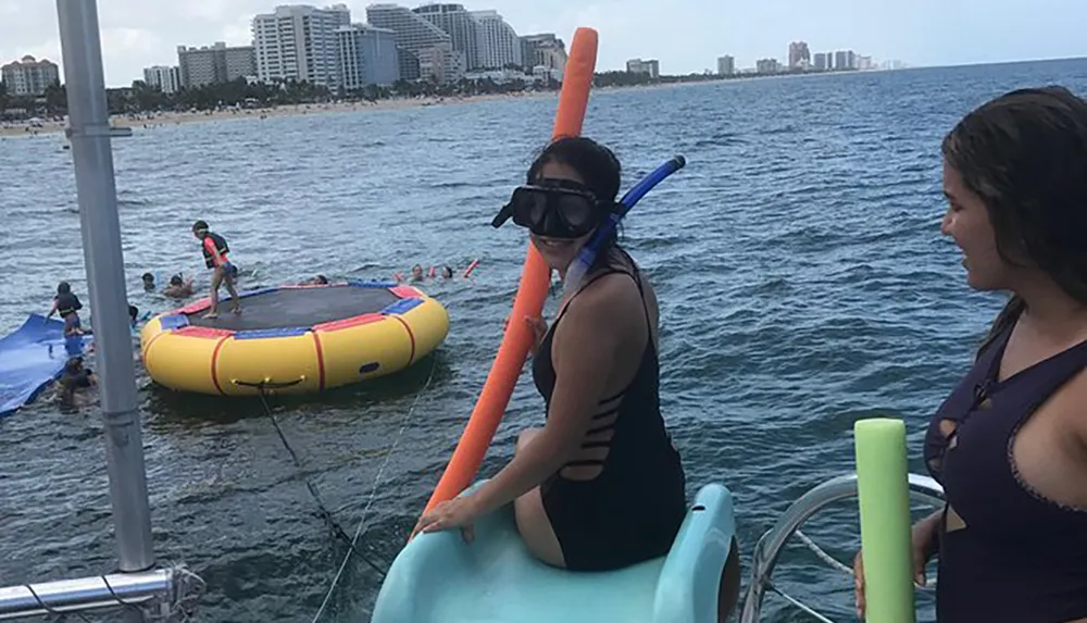 Two individuals are on a boat near a trampoline float in the water with one wearing a snorkel mask and holding a noodle against a backdrop of a coastal cityscape