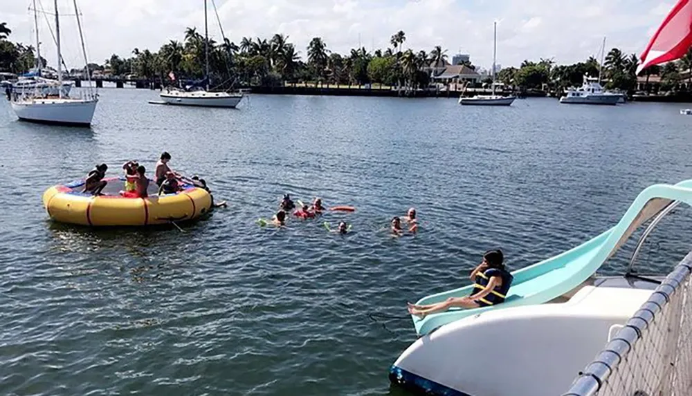 Children are enjoying a sunny day by playing in the water with an inflatable raft and a waterslide surrounded by boats in a marina