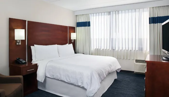 This is a neatly arranged hotel room with a large bed a side table a phone wall-mounted lights a chair and draped windows