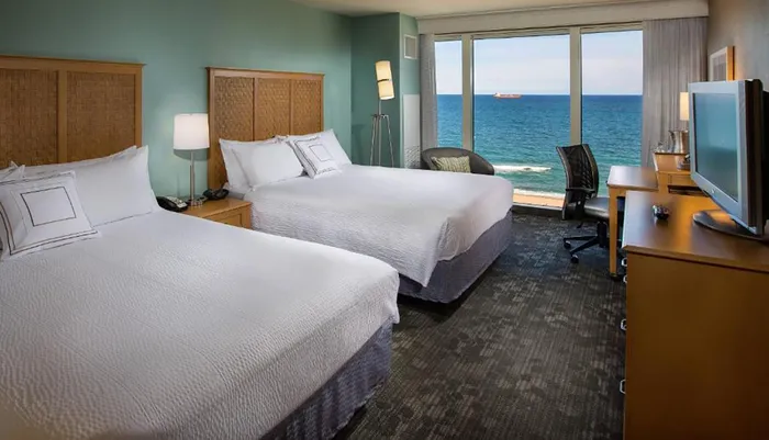 A hotel room with two beds features a large window offering a panoramic view of the ocean