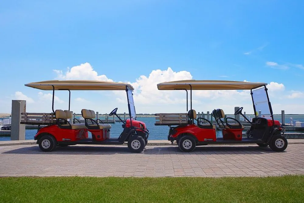 Two red golf carts are parked side by side on a paved area next to a body of water under a clear blue sky