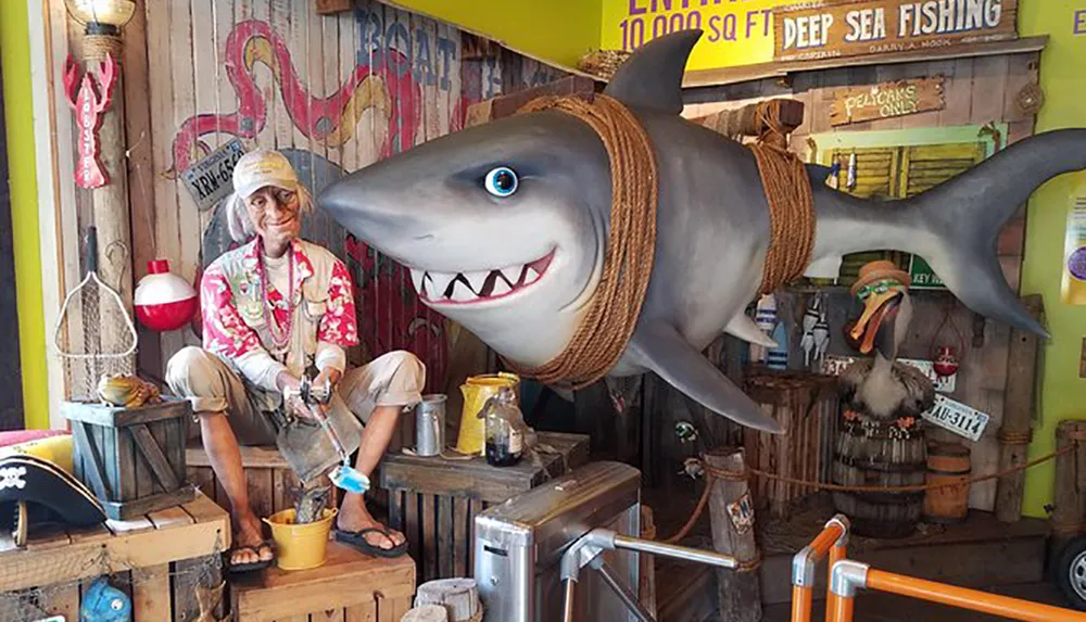 The image shows a whimsical display featuring a life-sized shark model wearing a hat alongside a mannequin of a fisherman set in a colorful nautical-themed environment