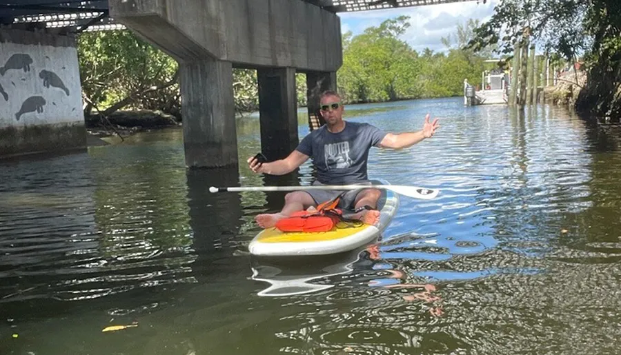 A person is relaxing on a paddleboard under a bridge, spreading his arms with a serene expression on his face.