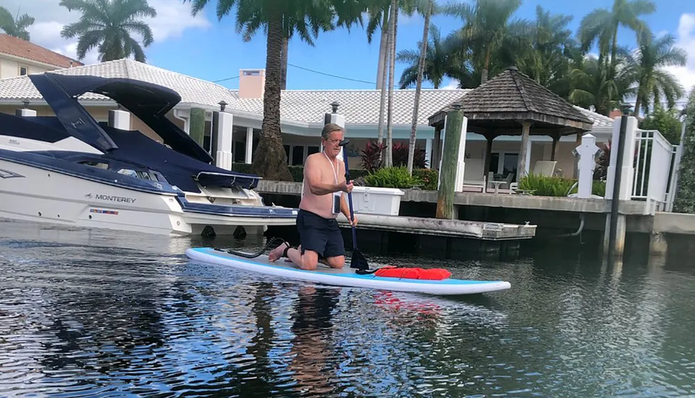 A person is kneeling on a paddleboard in a waterway with tropical surroundings and a yacht docked in the background