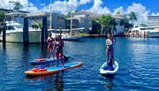 Three people are stand-up paddleboarding in a sunny waterway with boats and waterfront homes in the background.
