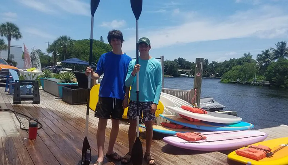 Two individuals are holding paddles on a sunny dock with colorful paddleboards and kayaks nearby ready for a water adventure