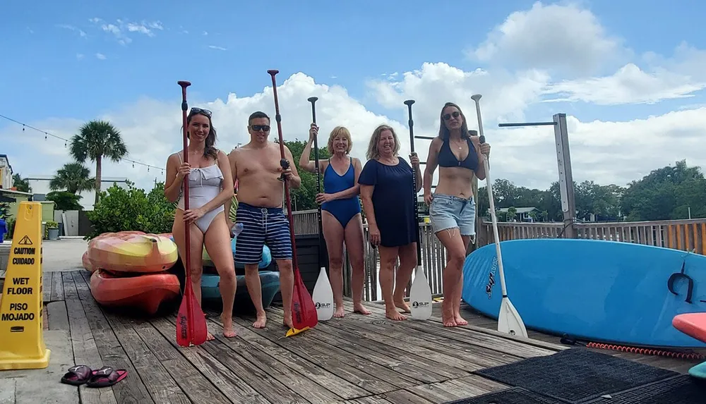 Five people in swimwear are standing on a wooden dock holding paddles ready for some water activities with kayaks and a paddleboard nearby under a partly cloudy sky
