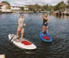Two people are paddleboarding on a calm river with waterfront houses in the background
