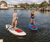 Two people are paddleboarding on a calm river with waterfront houses in the background