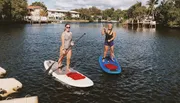 Two people are paddleboarding on a calm river with waterfront houses in the background.