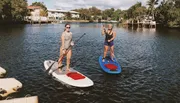 Two people are paddleboarding on a calm river with waterfront houses in the background.