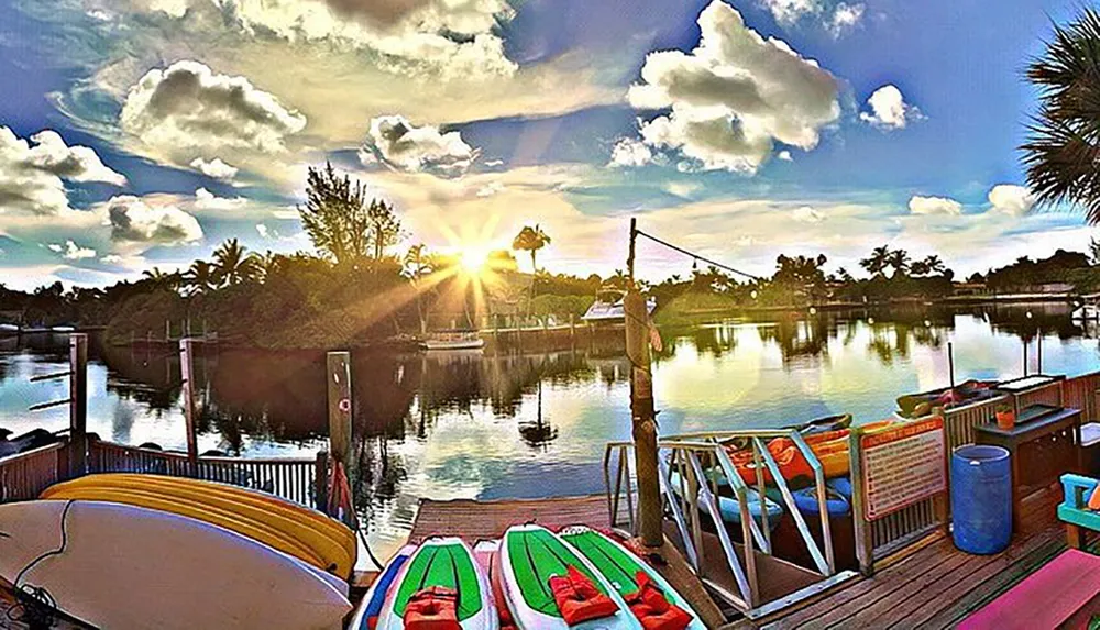 The image shows a vibrant sunset view over a calm waterway with colorful kayaks in the foreground and lush greenery under a dynamic sky