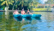 Three individuals are enjoying their time on a kayak in a scenic waterway surrounded by palm trees and waterfront properties.